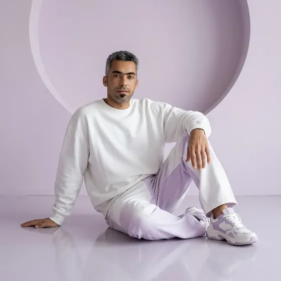 A man with short salt-and-pepper hair and a goatee sitting on the glossy floor of a pale lilac studio in an oversized white sweatshirt and white-and-lilac trousers
