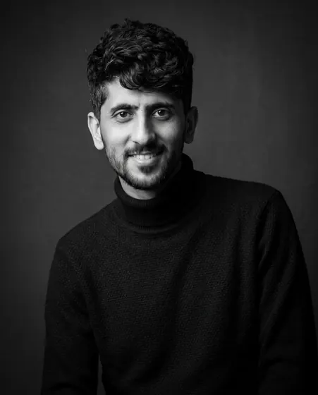 Black-and-white studio portrait of a young man with dark curly hair and a short beard in a black turtleneck on a charcoal backdrop, edited from a real photo