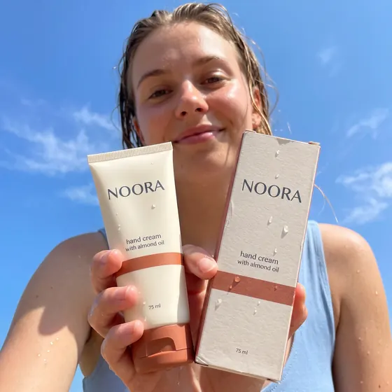 A cream-white hand-cream tube with a terracotta cap and its box held toward the camera from a low angle by a smiling young woman against a blue sky